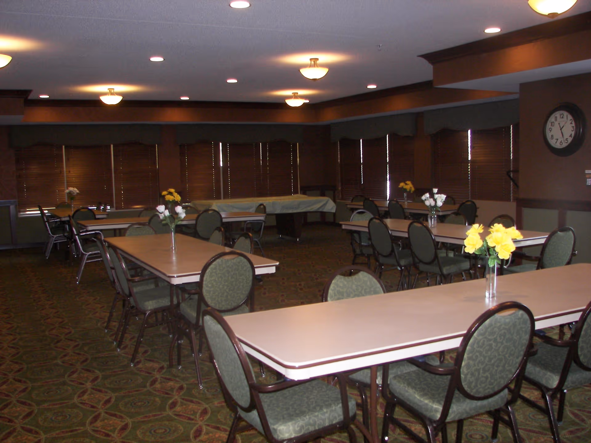 A dining room with several rectangular tables and green cushioned chairs arranged around them. Each table has a small vase with yellow or white flowers. The room has brown walls, a patterned carpet, multiple ceiling lights, and windows with closed blinds. A clock is mounted on one wall.