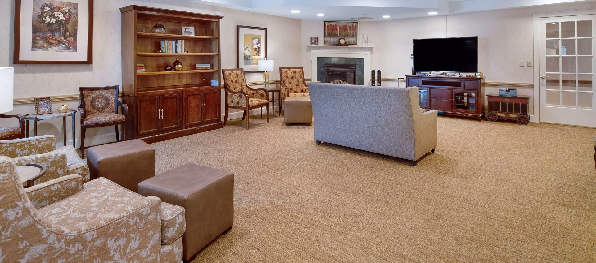 Spacious senior lounge with upholstered chairs and ottomans, a sofa facing a TV on a media console, a fireplace, and bookshelves.