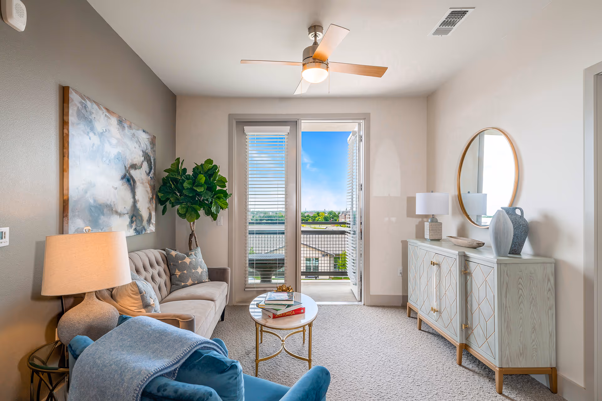 A bright and cozy living room with a beige tufted sofa, a blue armchair with a blue throw blanket, a round coffee table with books, a sideboard with decorative items and a round mirror above it, a large abstract painting on the wall, a potted plant, and a ceiling fan. French doors open to a balcony with a view of rooftops and greenery under a blue sky.