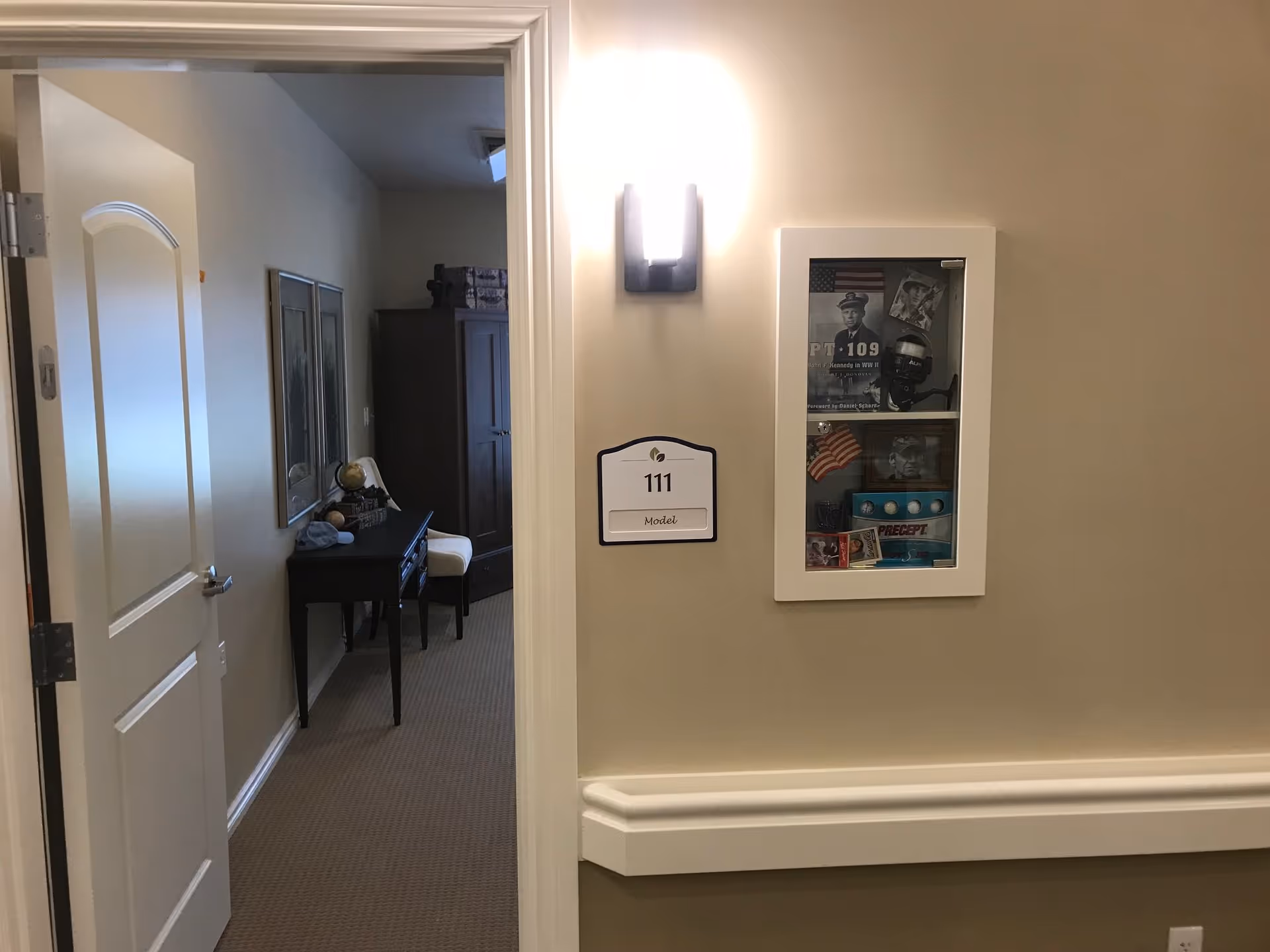 View of an open doorway leading into a room with a desk, chair, and wardrobe. The hallway wall outside the room has a light fixture, a room number sign labeled '111 Model', and a display case with memorabilia including a photo of a man in uniform, an American flag, and other vintage items.