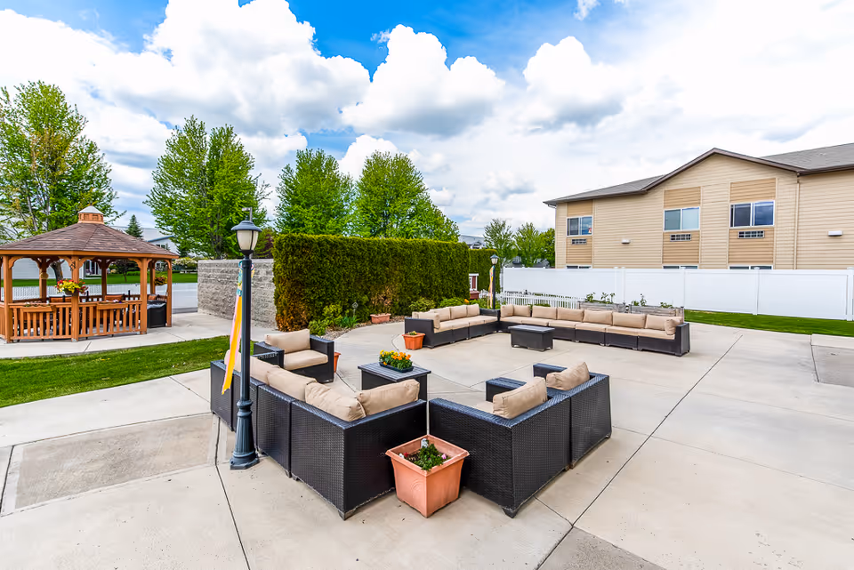 Outdoor patio area at Ellensburg Senior Living featuring a large square arrangement of cushioned wicker seating around a central table, with potted plants and a wooden gazebo in the background, surrounded by green trees and a beige building under a partly cloudy blue sky.