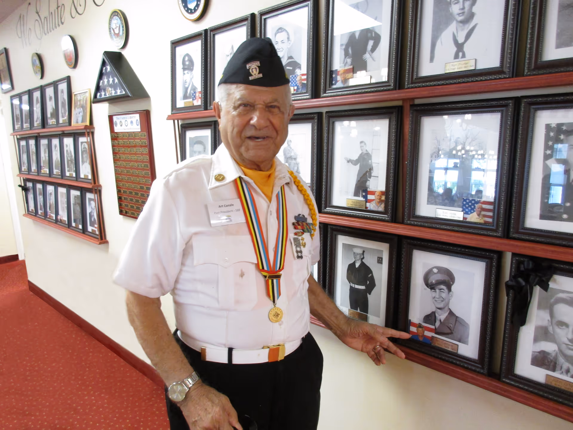 An elderly man wearing a white military uniform with medals and a black beret stands in a hallway with red carpet. He is pointing to a framed black and white photograph of a serviceman on the wall. The wall is decorated with multiple framed photographs of military personnel and a folded American flag display case.