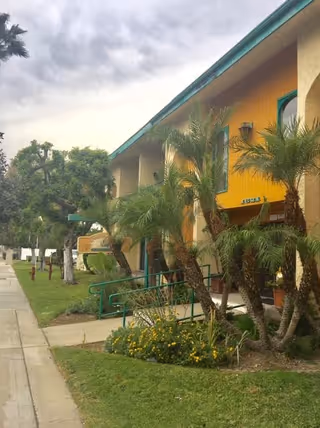 Exterior view of Cambridge Court Assisted Living facility showing a two-story building with a yellow and beige facade, green trim, palm trees, and a sidewalk with grass and flowers alongside it under a cloudy sky.