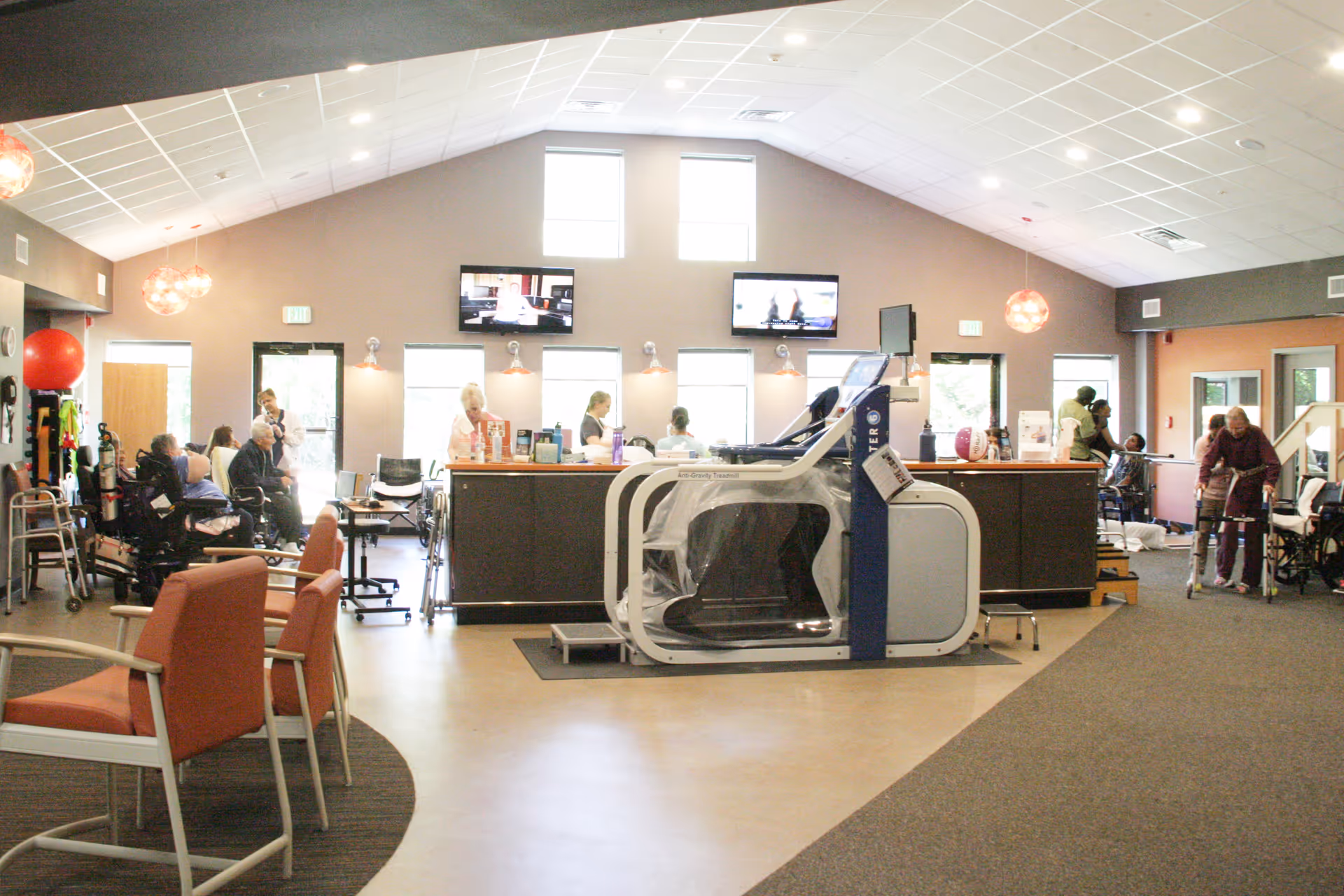 A spacious senior living facility common area with several elderly residents and staff. The room features a large anti-gravity treadmill in the center, multiple chairs, walkers, and wheelchairs. There are three wall-mounted televisions above a counter where staff are assisting residents. The room has high ceilings with hanging lights and large windows letting in natural light.