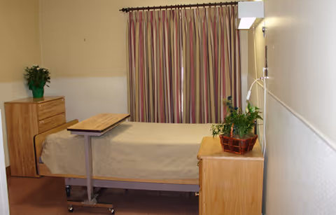 A simple healthcare facility bedroom with a single bed covered in a beige blanket, a wooden bedside table with a potted plant, a wooden dresser with another potted plant, and striped curtains covering the window.
