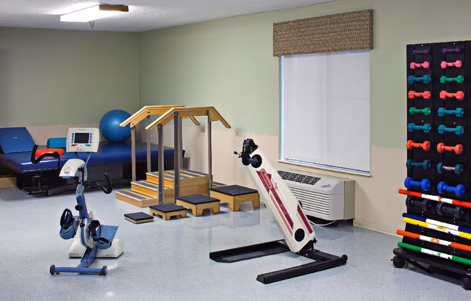 A physical therapy or exercise room with equipment including a stationary bike, a set of wooden stairs with handrails, a blue exercise ball, a padded therapy table, a leg exercise machine, and a rack holding colorful dumbbells and exercise bars. The room has a window with a closed blind and an air conditioning unit below it.