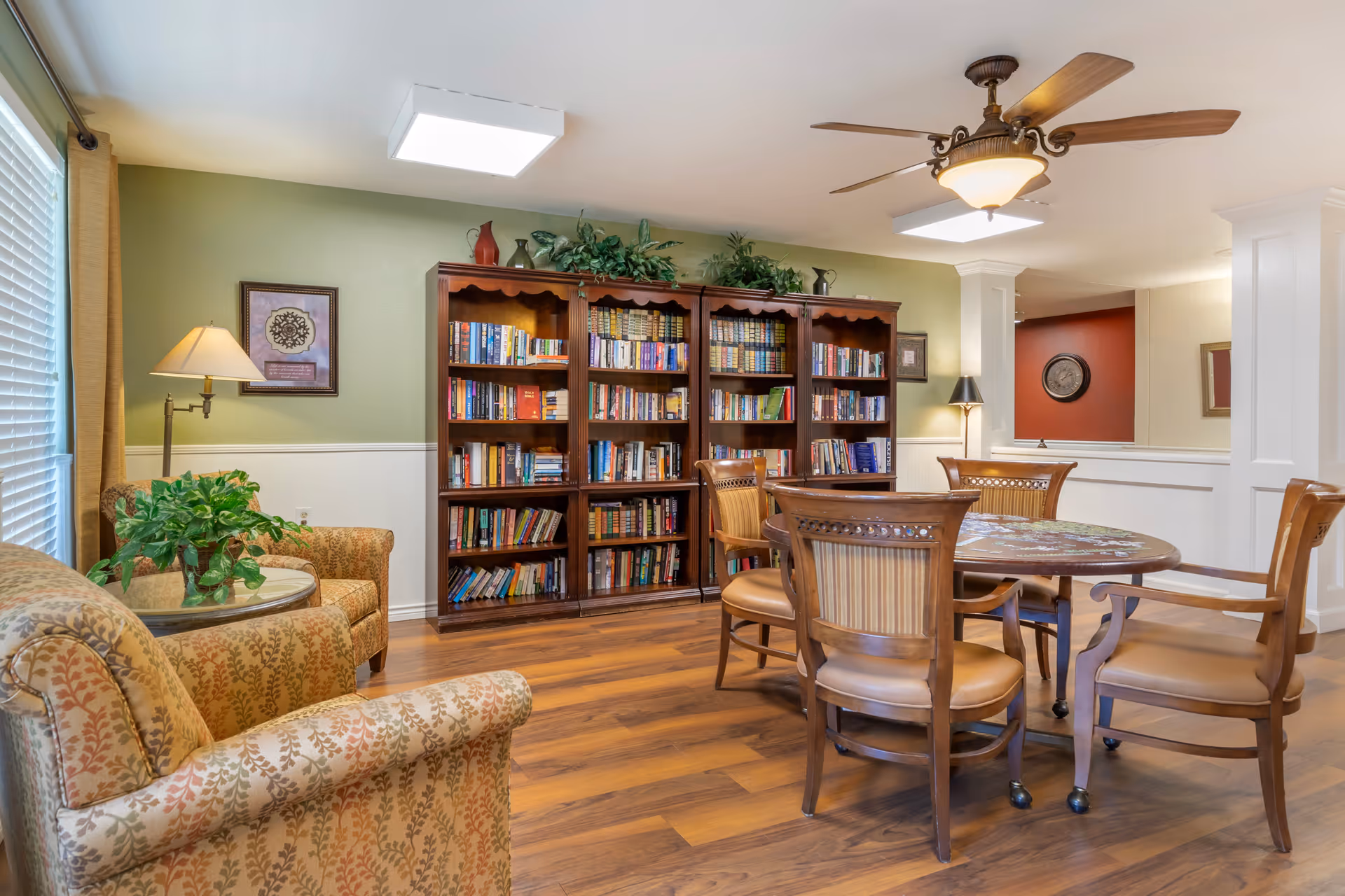 A cozy reading and sitting area in a senior living facility with a large wooden bookshelf filled with books against a green wall. There are four wooden chairs around a round table with a puzzle on it. Two upholstered armchairs with patterned fabric are placed near a window with blinds and beige curtains. The room has wooden flooring, a ceiling fan with a light, and two standing lamps providing warm lighting.