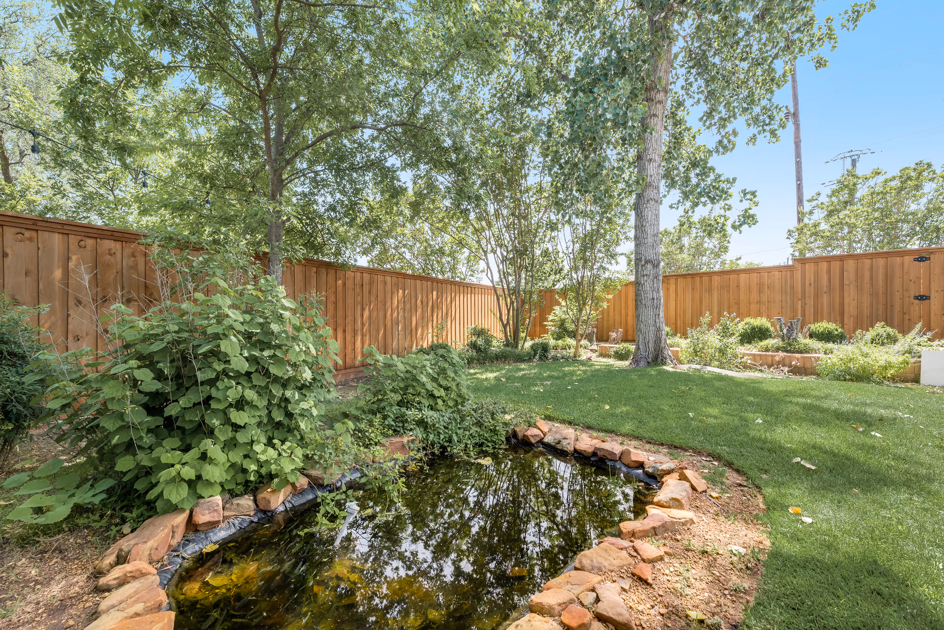 A peaceful outdoor garden area with a small pond surrounded by rocks, green grass, various bushes, and tall trees. The garden is enclosed by a wooden fence under a clear blue sky.