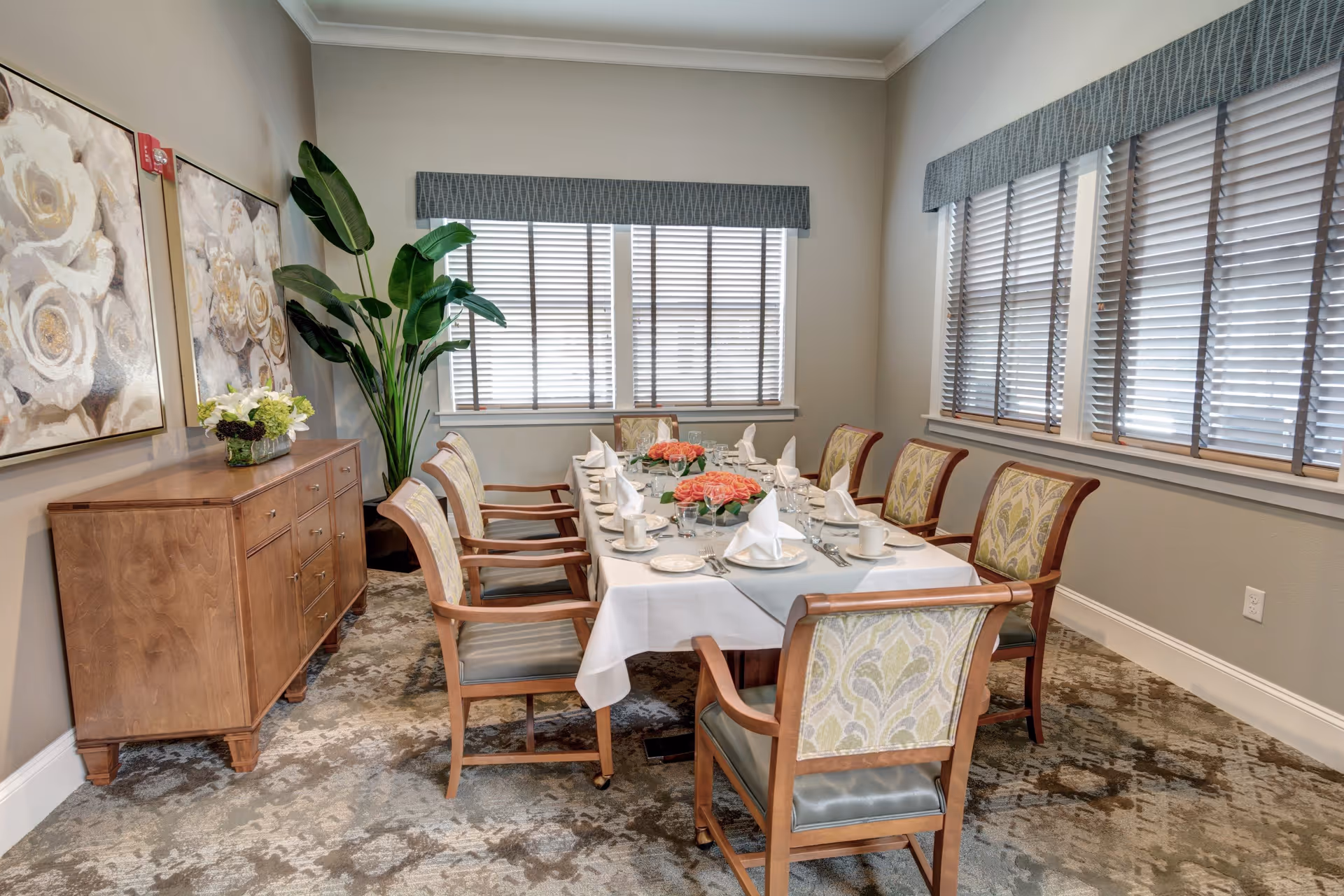 A dining room with a rectangular table set for eight people. The table is covered with a white tablecloth and has floral centerpieces with pink flowers. Each place setting includes a white napkin, plate, cup, and glassware. The room has two large windows with blinds and valances, a wooden sideboard with a flower arrangement, two large floral paintings on the wall, and a tall green plant in the corner. The floor is carpeted with a patterned design.