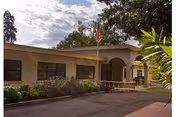 Exterior view of a single-story building with a covered entrance, an American flag on a flagpole, and surrounding greenery under a partly cloudy sky.