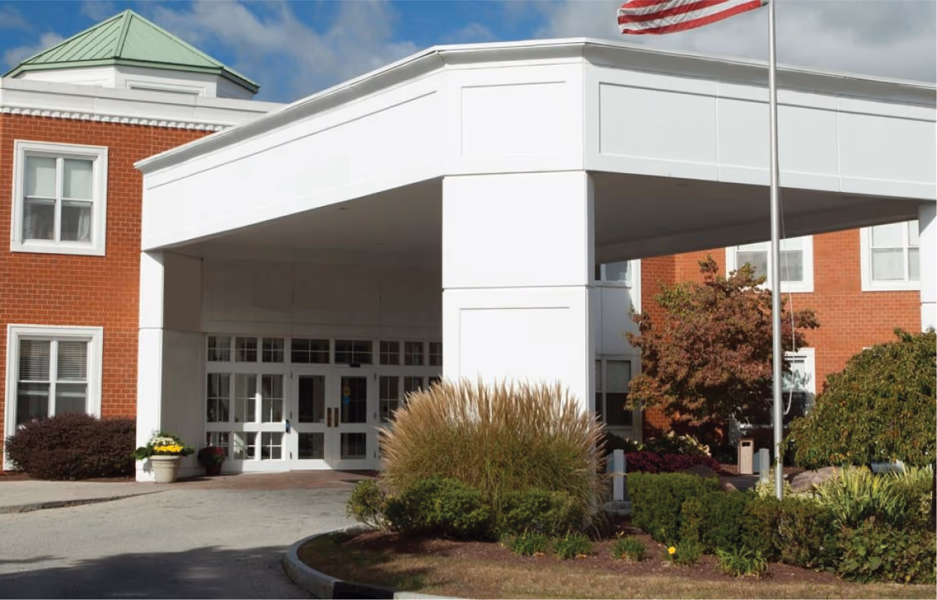 Entrance of Taconic Rehabilitation and Nursing at Hopewell facility showing a covered drop-off area with white columns, a brick building with white-framed windows, landscaped bushes and plants, and an American flag on a flagpole.