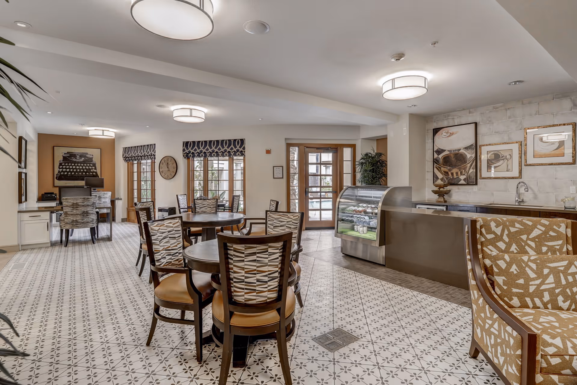 A bright and spacious common area in a senior living facility featuring round tables with patterned chairs, a small counter with a glass display case containing food items, framed artwork on the walls, large windows with patterned valances, and a desk with a computer in the background.