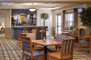 A cozy dining area in a senior living facility featuring wooden tables and chairs with cushioned seats. The room has a carpeted floor with a patterned design, a bar counter with high chairs, and large windows with blinds allowing natural light. The walls are painted in neutral tones, and there are plants and decorative elements adding warmth to the space.