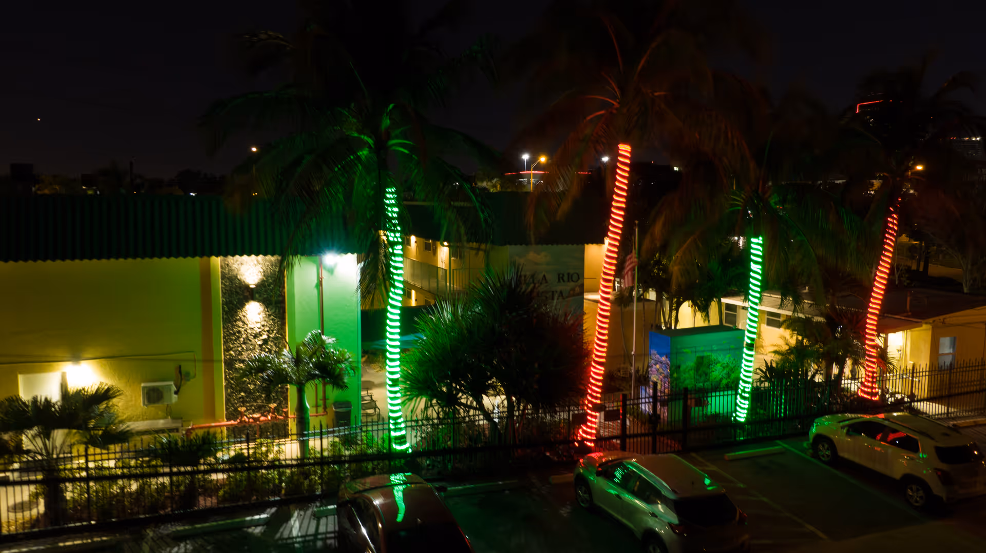 Night view of Villa Rio Vista facility with palm trees wrapped in red and green lights, a yellow building in the background, and parked cars in the foreground.