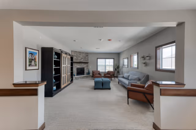 A spacious living room with beige carpet, a gray sofa, two brown leather armchairs, and two blue ottomans. There is a stone fireplace at the far end of the room, flanked by windows letting in natural light. A large black shelving unit is on the left wall, and framed artwork and small plants decorate the walls.