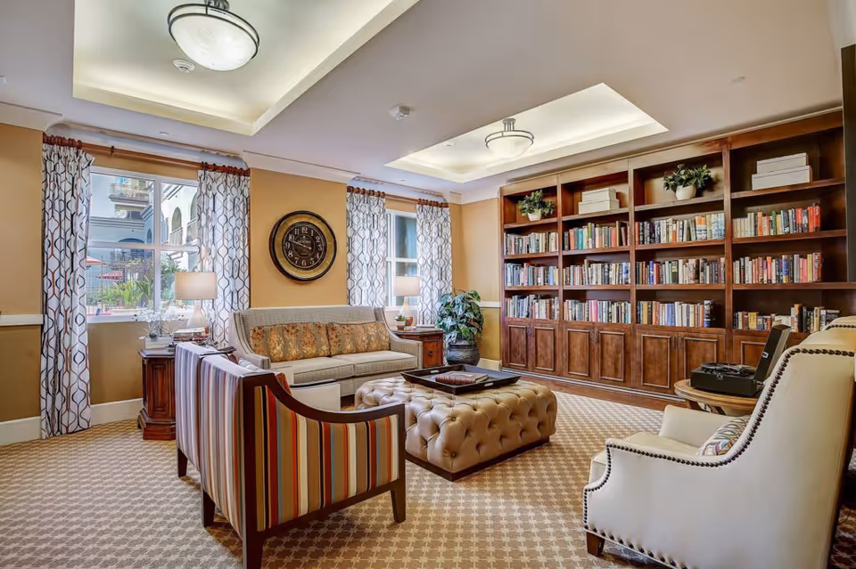 A cozy living room area with a large wooden bookshelf filled with books, a beige tufted ottoman in the center, a striped armchair, a beige sofa with patterned cushions, and a white armchair with nailhead trim. The room has two windows with patterned curtains, a round wall clock, two table lamps on wooden side tables, and a patterned carpet.