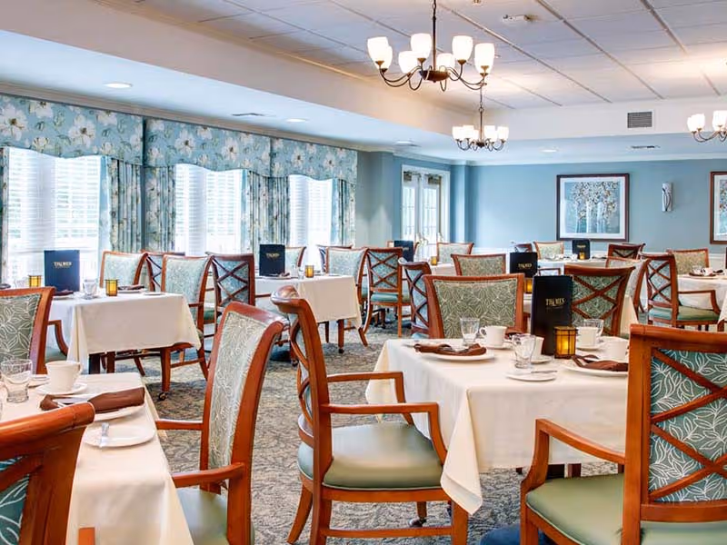 A bright and elegant dining room with multiple tables covered in white tablecloths, set with cups, glasses, plates, and brown napkins. The chairs have wooden frames with green patterned upholstery. Large windows with floral valances allow natural light to fill the room. Chandeliers hang from the ceiling, and framed artwork decorates the light blue walls.