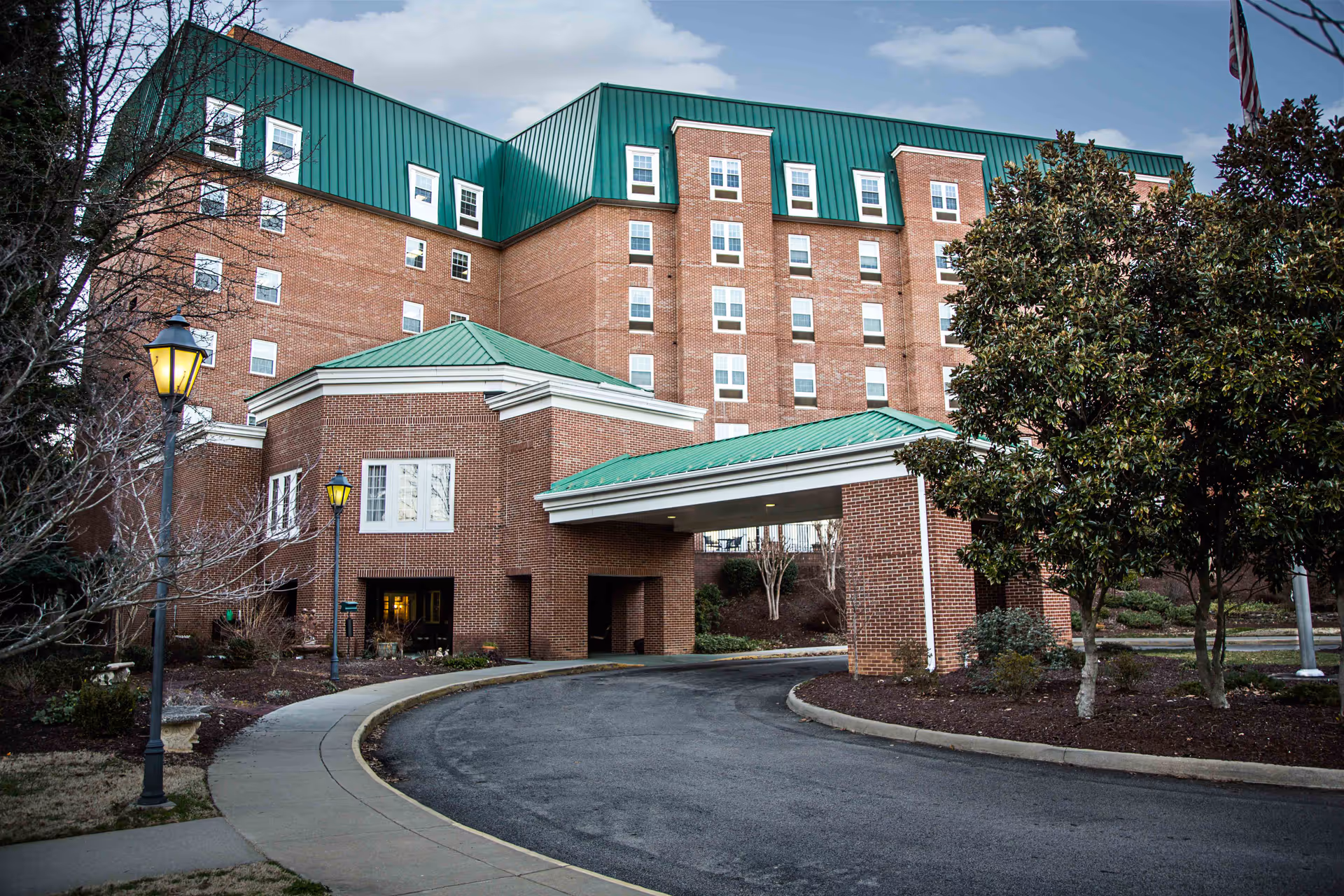 Exterior view of a multi-story brick building with a green metal roof, featuring a covered driveway entrance. The surrounding area includes a curved driveway, street lamps, trees, and landscaped bushes under a partly cloudy sky.