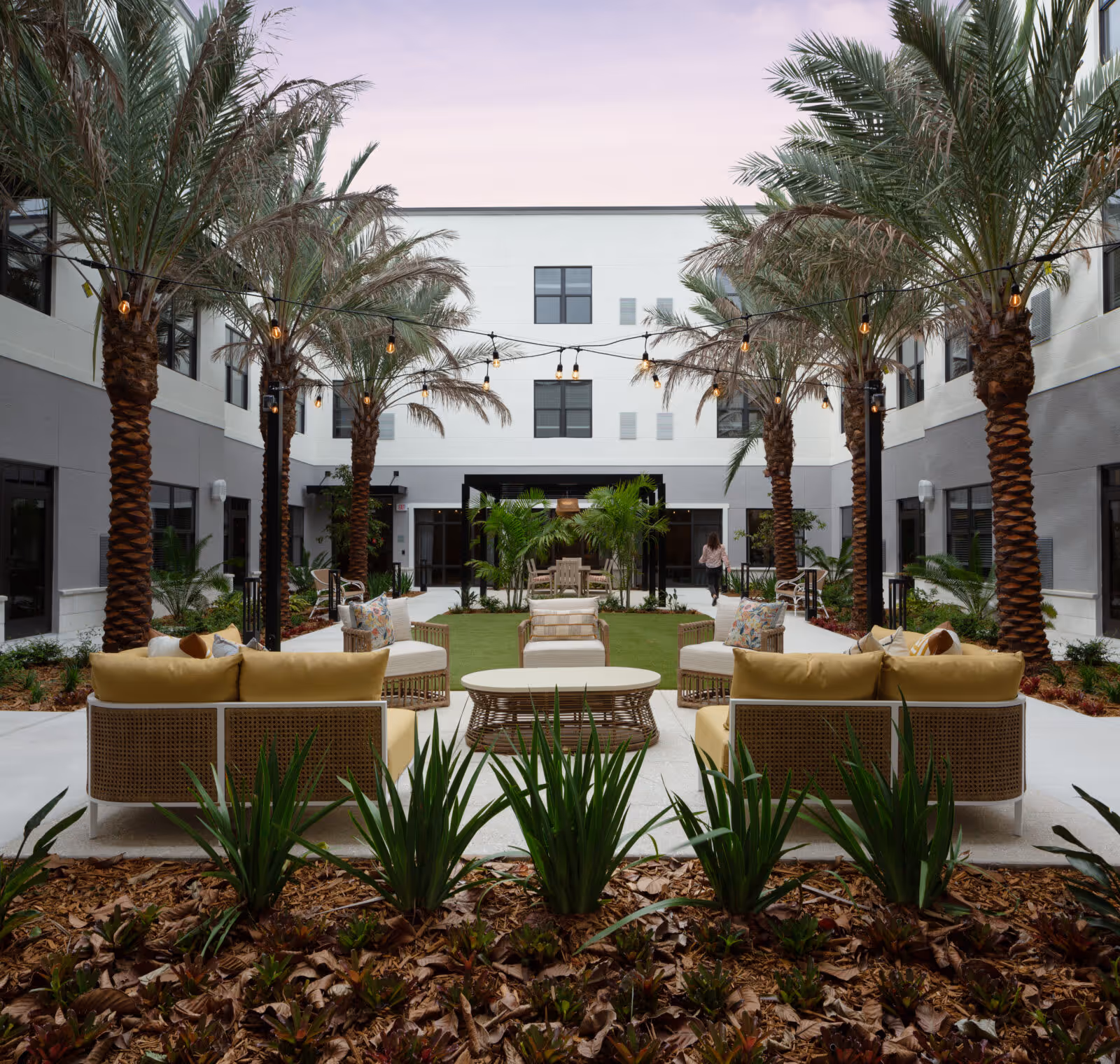 Outdoor courtyard area at The Palms at Plantation featuring palm trees, string lights, cushioned seating with yellow pillows, a central coffee table, and landscaped plants with a building surrounding the courtyard.