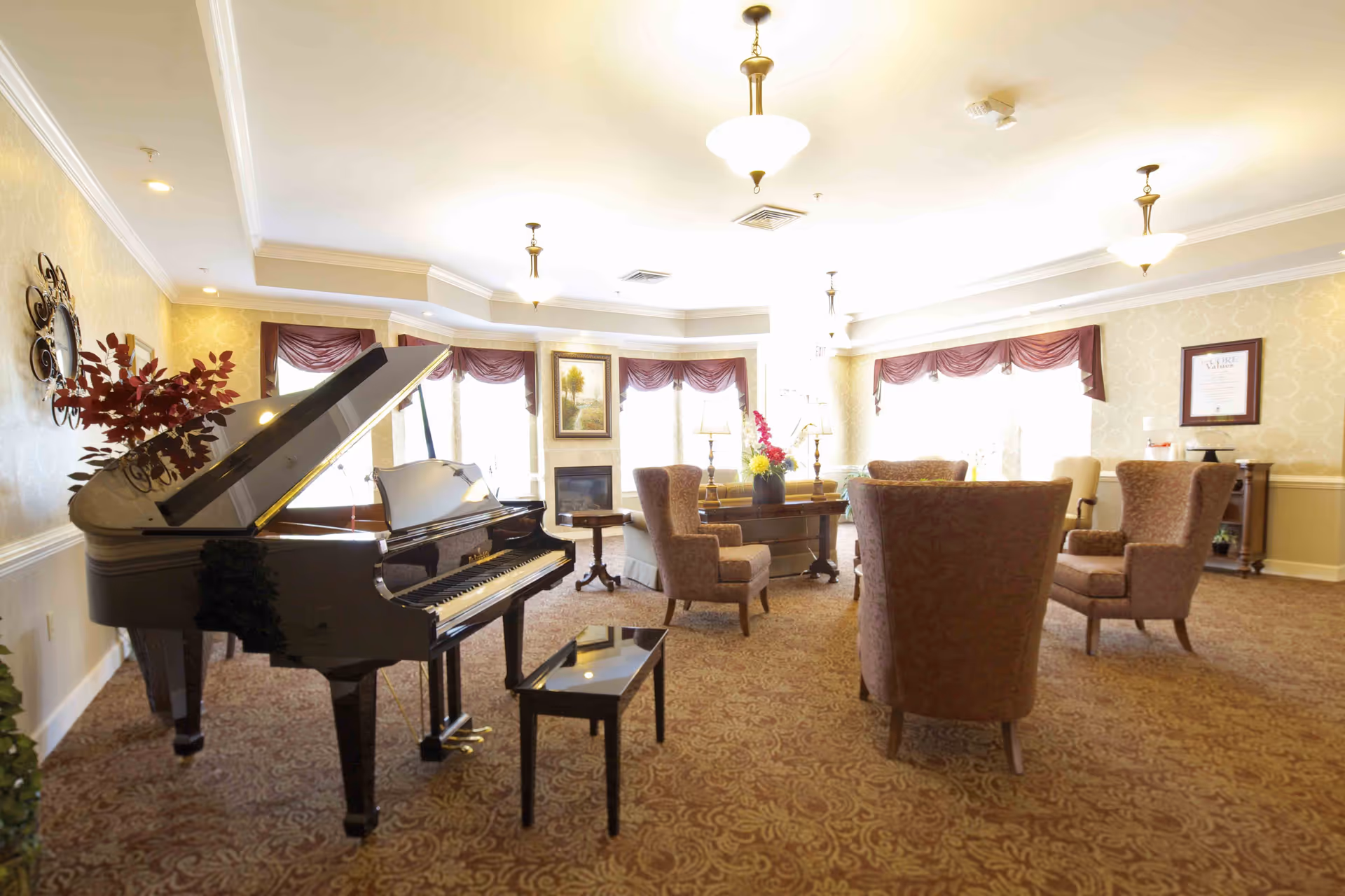 A spacious senior living common area with a grand piano on the left, several upholstered armchairs arranged around a central table with a floral arrangement, large windows with burgundy valances, and warm lighting from ceiling fixtures.