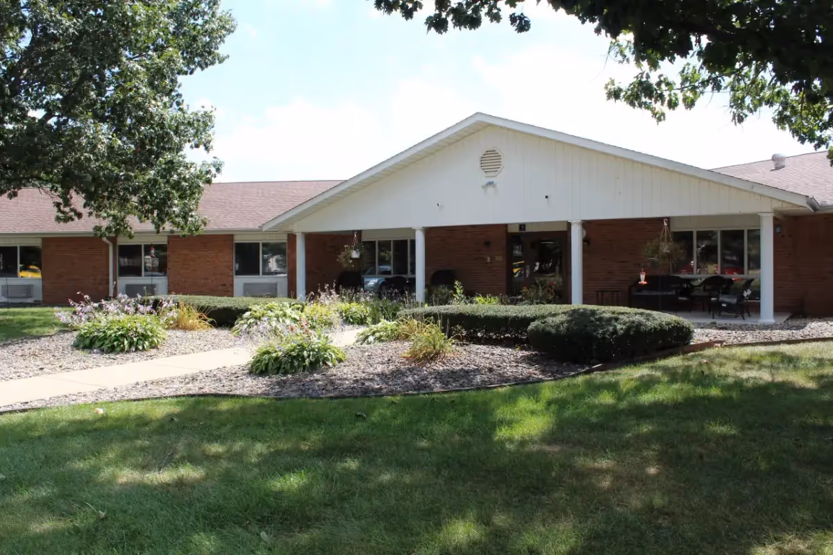 Front exterior view of a single-story brick building with a white gabled roof and a covered porch area. There are shrubs, flower beds, and a walkway leading up to the entrance. Trees provide some shade over the grassy lawn in the foreground.