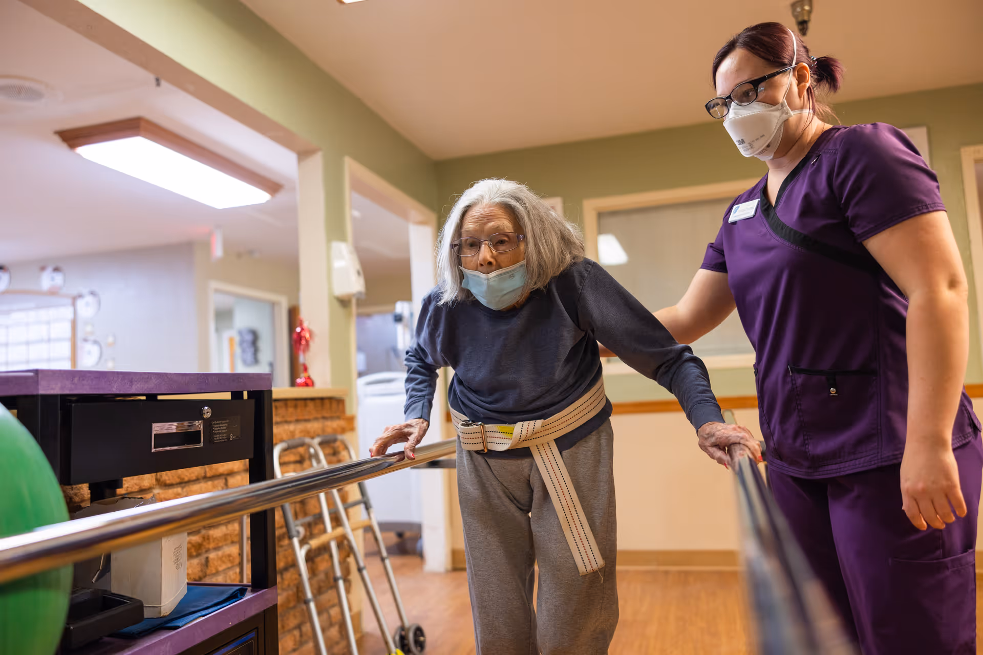 An elderly woman wearing a face mask and glasses is using parallel bars for physical therapy while being assisted by a healthcare worker in purple scrubs and a face mask inside a rehabilitation center.