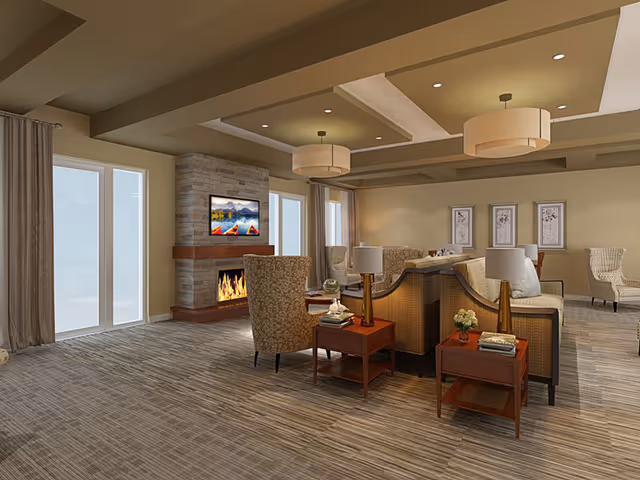 A spacious and well-lit living room area with beige walls and ceiling featuring recessed lighting and two large round pendant lights. The room has a stone fireplace with a flat-screen TV mounted above it, showing an image of colorful kayaks on a lake with mountains in the background. There are multiple comfortable armchairs and sofas arranged around wooden side tables with lamps and decorative items. Large windows with curtains allow natural light into the room, and framed artwork is displayed on the far wall.