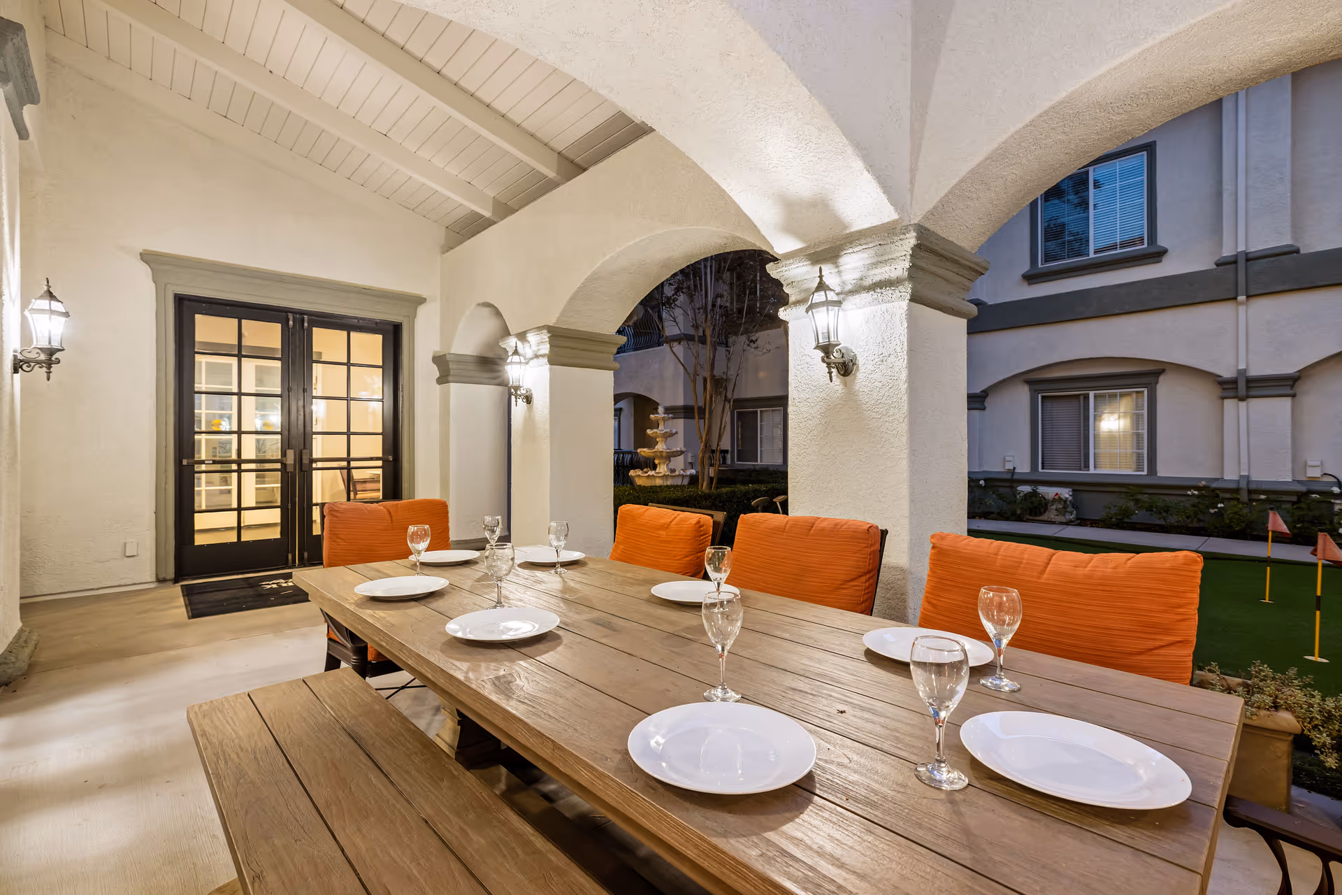 Outdoor covered patio area with a wooden dining table set with white plates and wine glasses. The table has orange cushioned chairs and a wooden bench. The patio features white stucco arches and wall-mounted lantern lights. In the background, there is a courtyard with a fountain and a putting green with small flags.