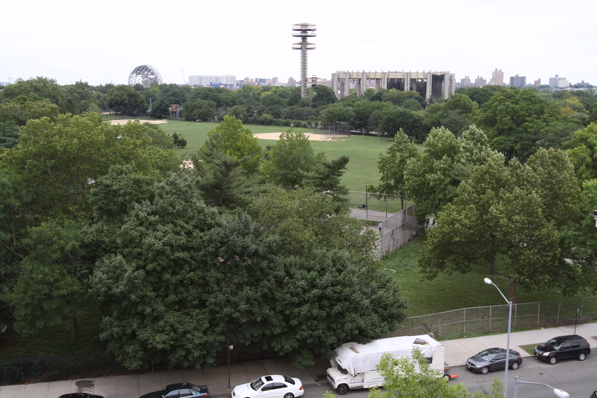 View from above of a tree-lined park with baseball diamonds and a fenced sports court, bordered by a street with parked cars and a white covered truck.