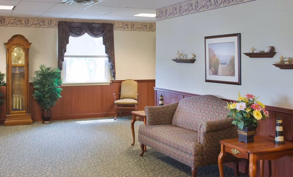 A cozy seating area with a patterned loveseat, side table with flowers, a grandfather clock, and framed artwork in a senior center.