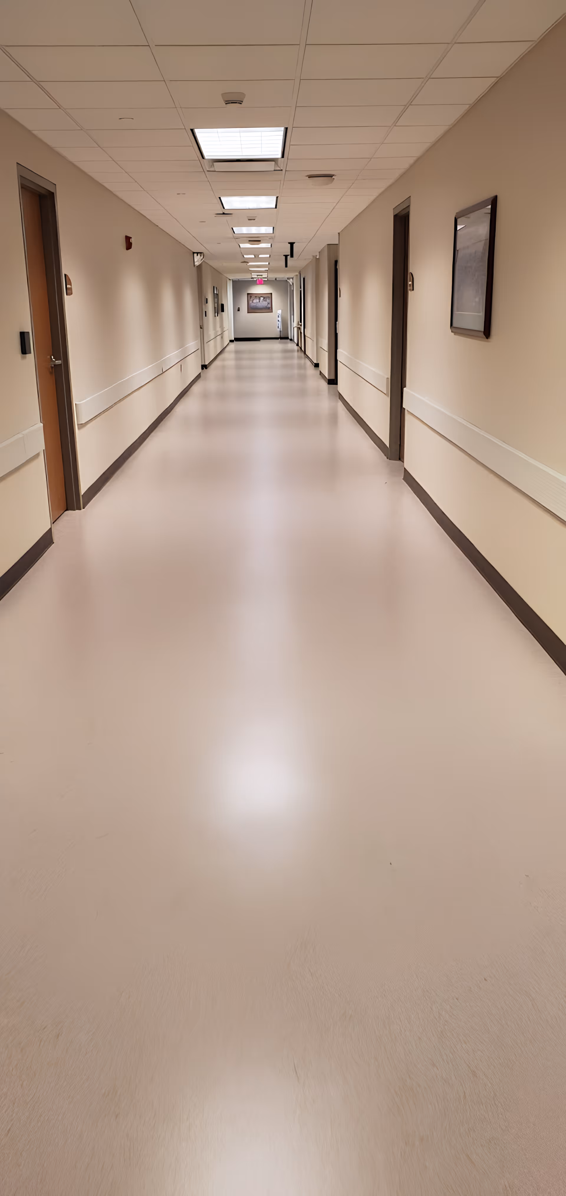 A long, empty hospital or medical facility hallway with beige walls, several closed doors on both sides, handrails along the walls, ceiling lights, and a framed picture at the far end.