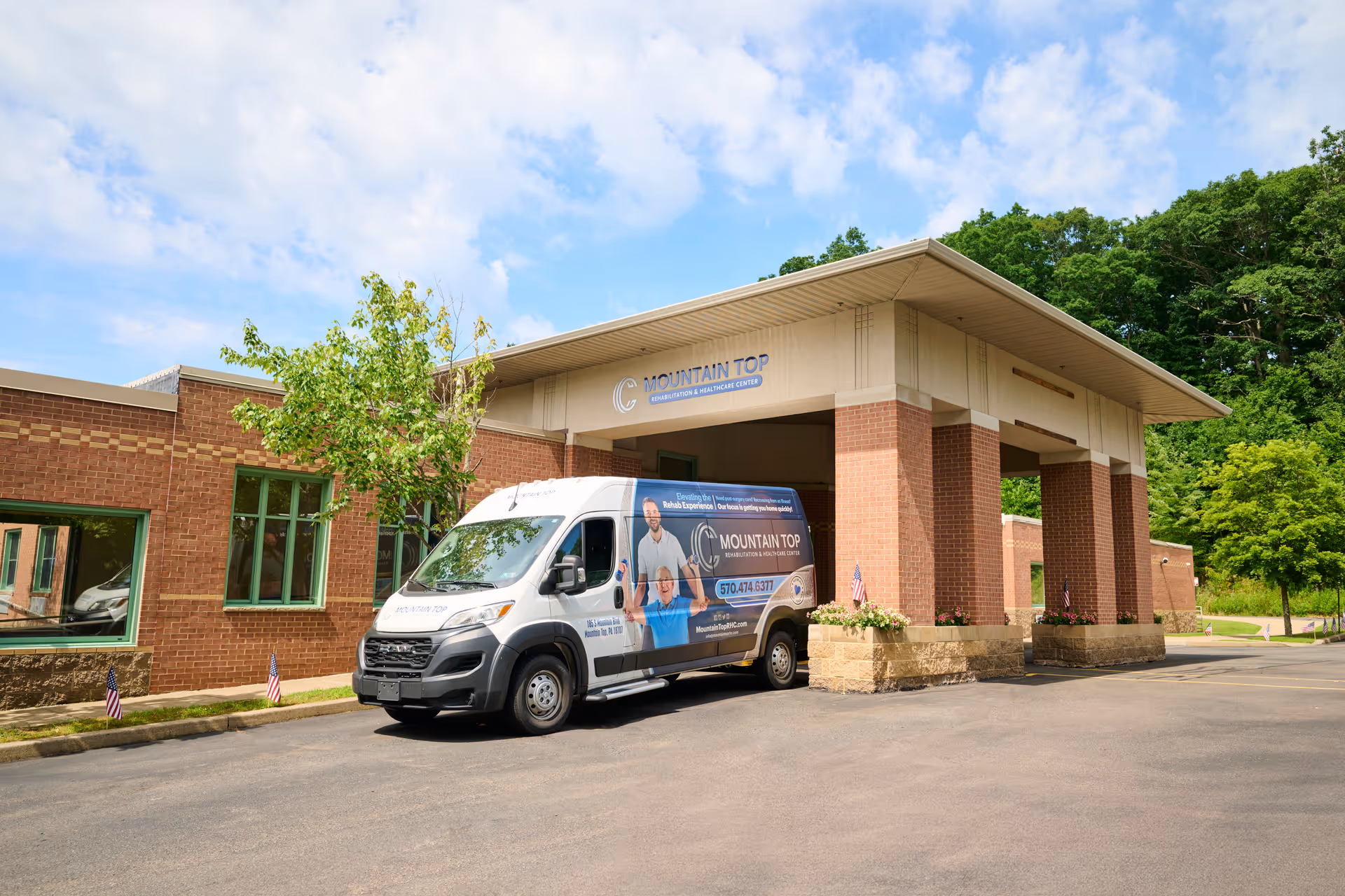 Exterior view of Mountain Top Rehabilitation and Healthcare Center showing the entrance with a covered drop-off area. A branded van with the facility's logo and contact information is parked in front. The building is made of brick with green-trimmed windows, and there are trees and a partly cloudy sky in the background.