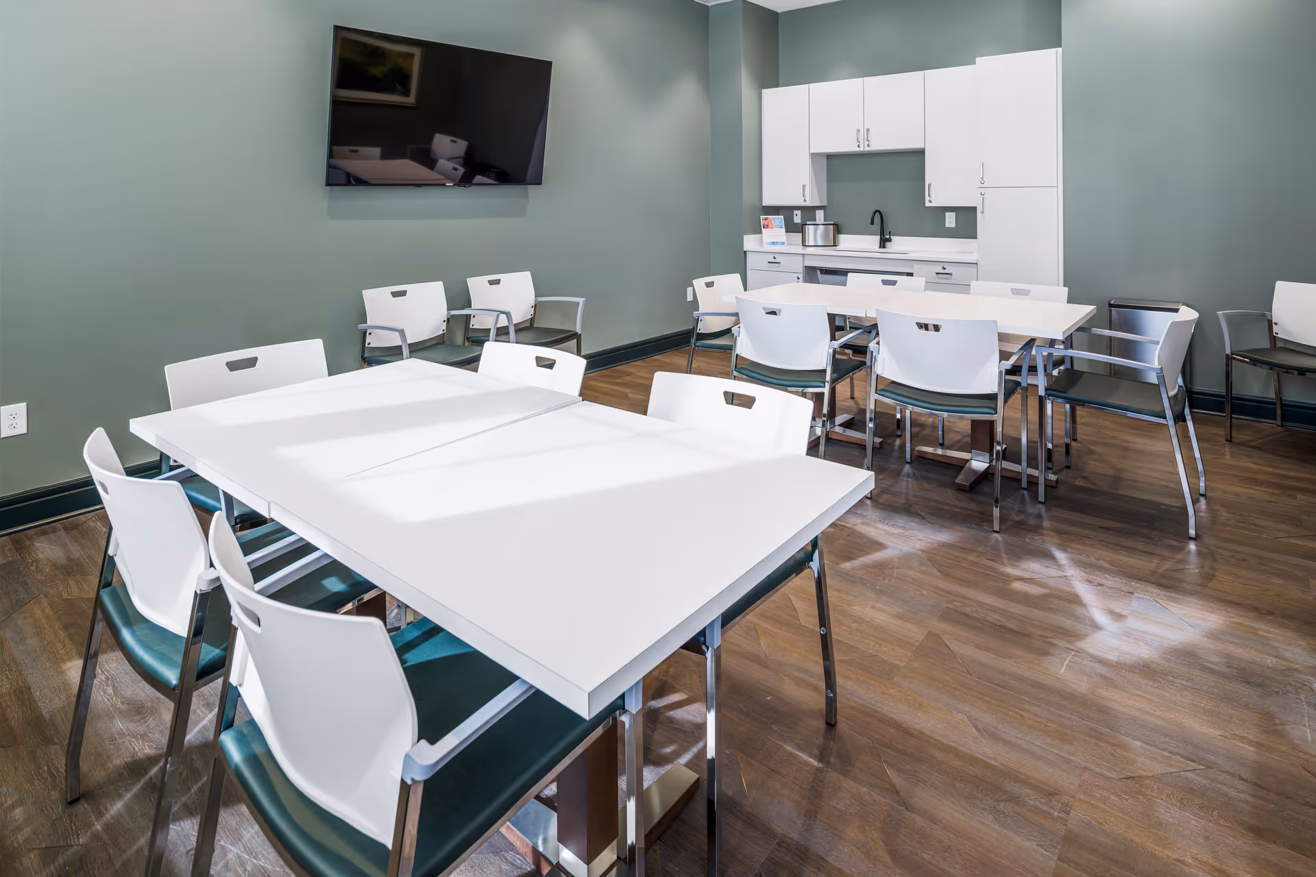 A clean and modern common area with white tables and chairs arranged on a wooden floor. The room has green walls, a mounted flat-screen TV, and a kitchenette with white cabinets, a sink, and a coffee maker.