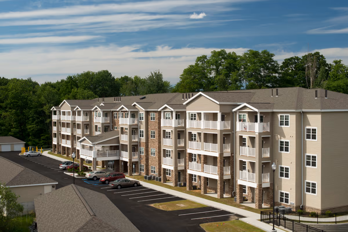 Exterior view of a multi-story residential building with balconies, a parking lot, and surrounding trees.