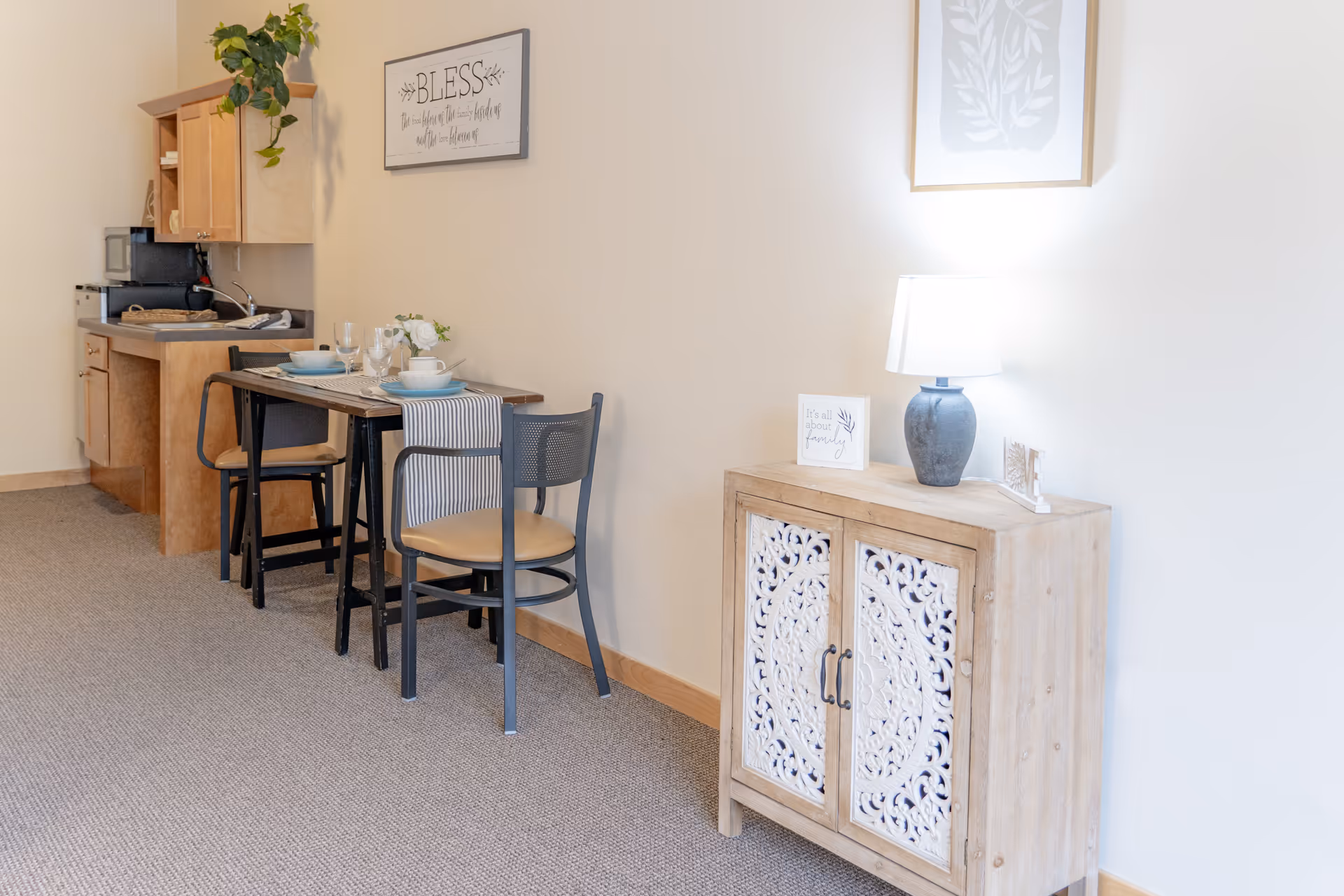 A small dining area with a wooden table set for two, featuring plates, bowls, glasses, and a striped table runner. Behind the table is a compact kitchenette with wooden cabinets, a microwave, and a sink. To the right, there is a decorative wooden cabinet with intricate white carvings, topped with a lamp and small framed signs. The walls are light-colored with framed artwork and a sign that reads 'BLESS'.