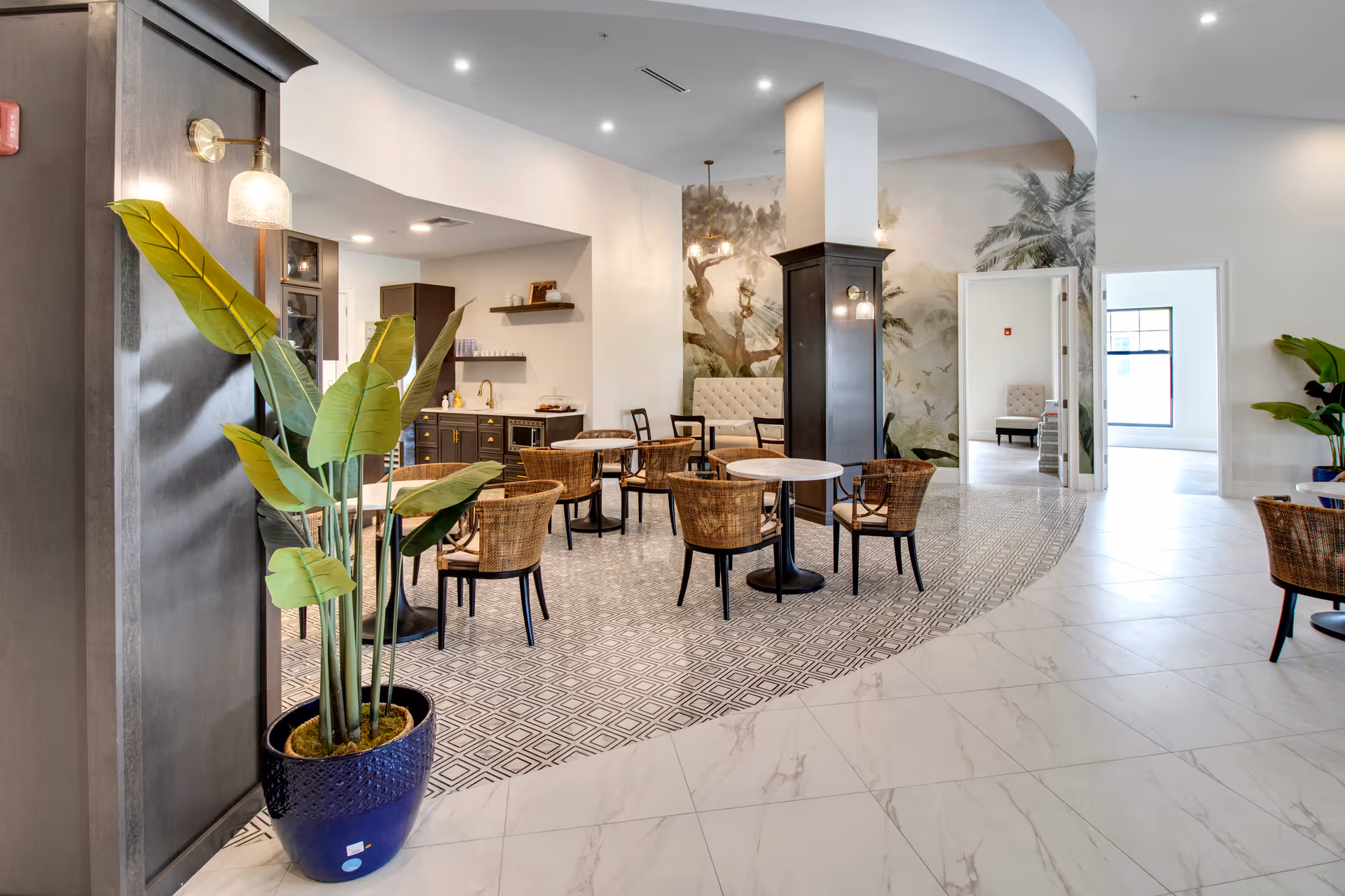 A bright and modern common area in an assisted living facility featuring a small kitchenette with dark cabinets, round tables with wicker chairs, a large potted plant in a blue pot, and a mural of trees and birds on the wall. The floor transitions from patterned tile to white marble-like tile, and there are open doorways leading to other rooms.