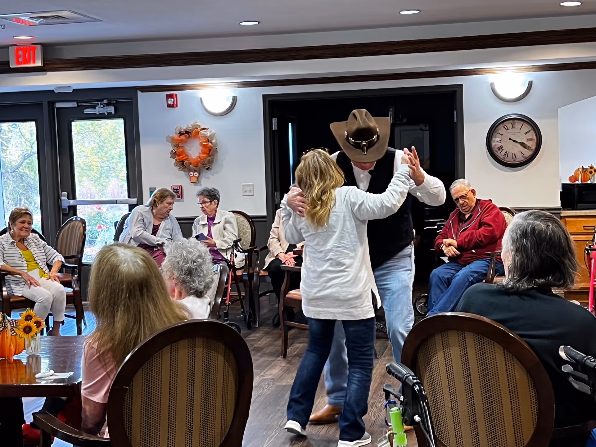A group of elderly people seated in a common room with wooden floors and chairs. In the center, a man wearing a cowboy hat and a woman in a white sweater are dancing. The room has a clock on the wall, a wreath decoration, and large windows letting in natural light.