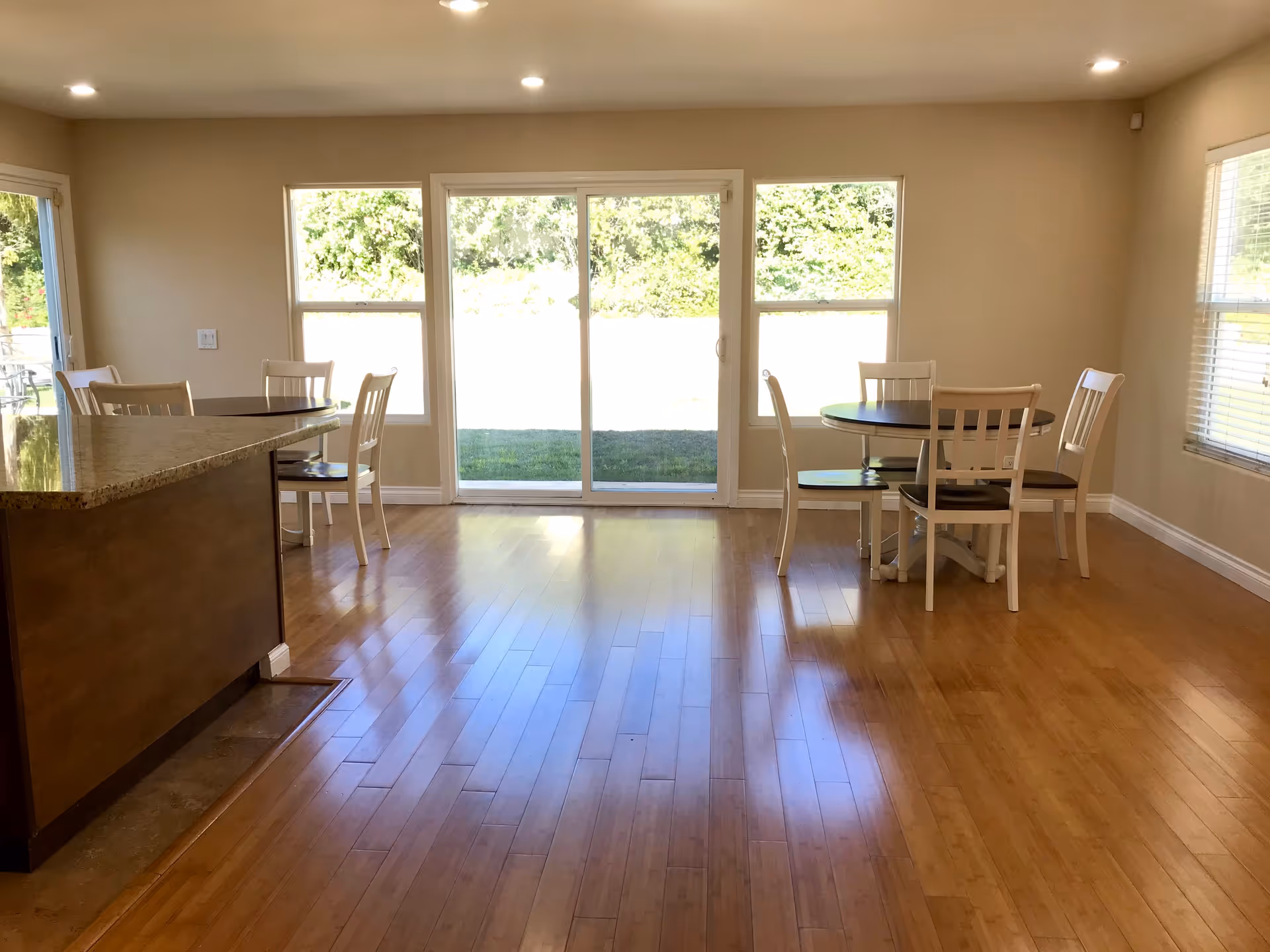 A bright dining area with wooden floors, two round tables each surrounded by four white chairs with dark seats, a kitchen counter with a granite countertop on the left, and large sliding glass doors and windows letting in natural light and showing greenery outside.