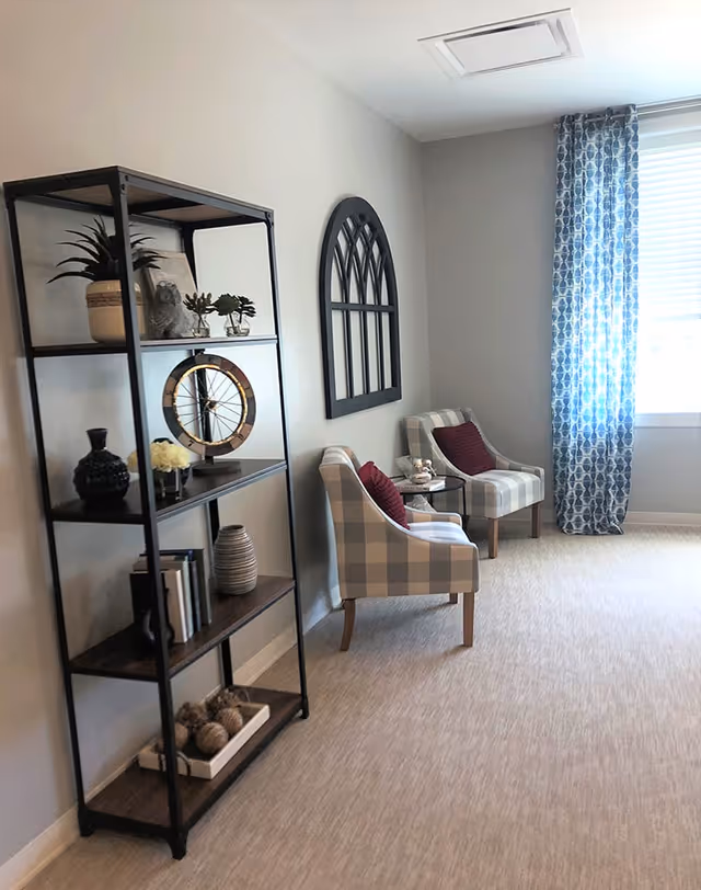 A cozy seating area with two plaid armchairs and a shelving unit decorated with vases and decor beside a window with a patterned curtain.