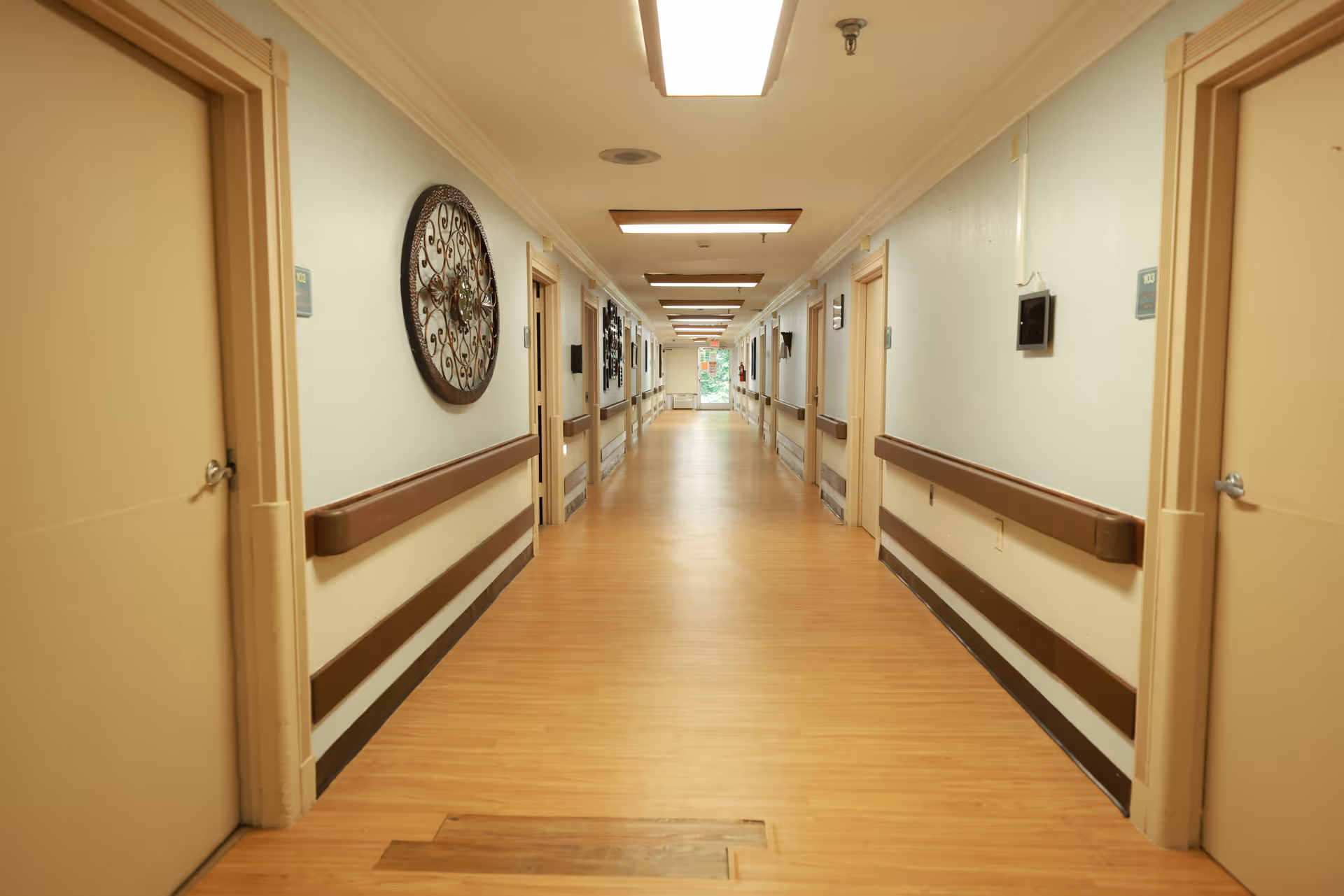 A long, well-lit hallway in a healthcare facility with beige walls and wooden flooring. Both sides of the hallway have multiple closed doors, handrails, and decorative wall art. The hallway extends towards a window at the far end letting in natural light.
