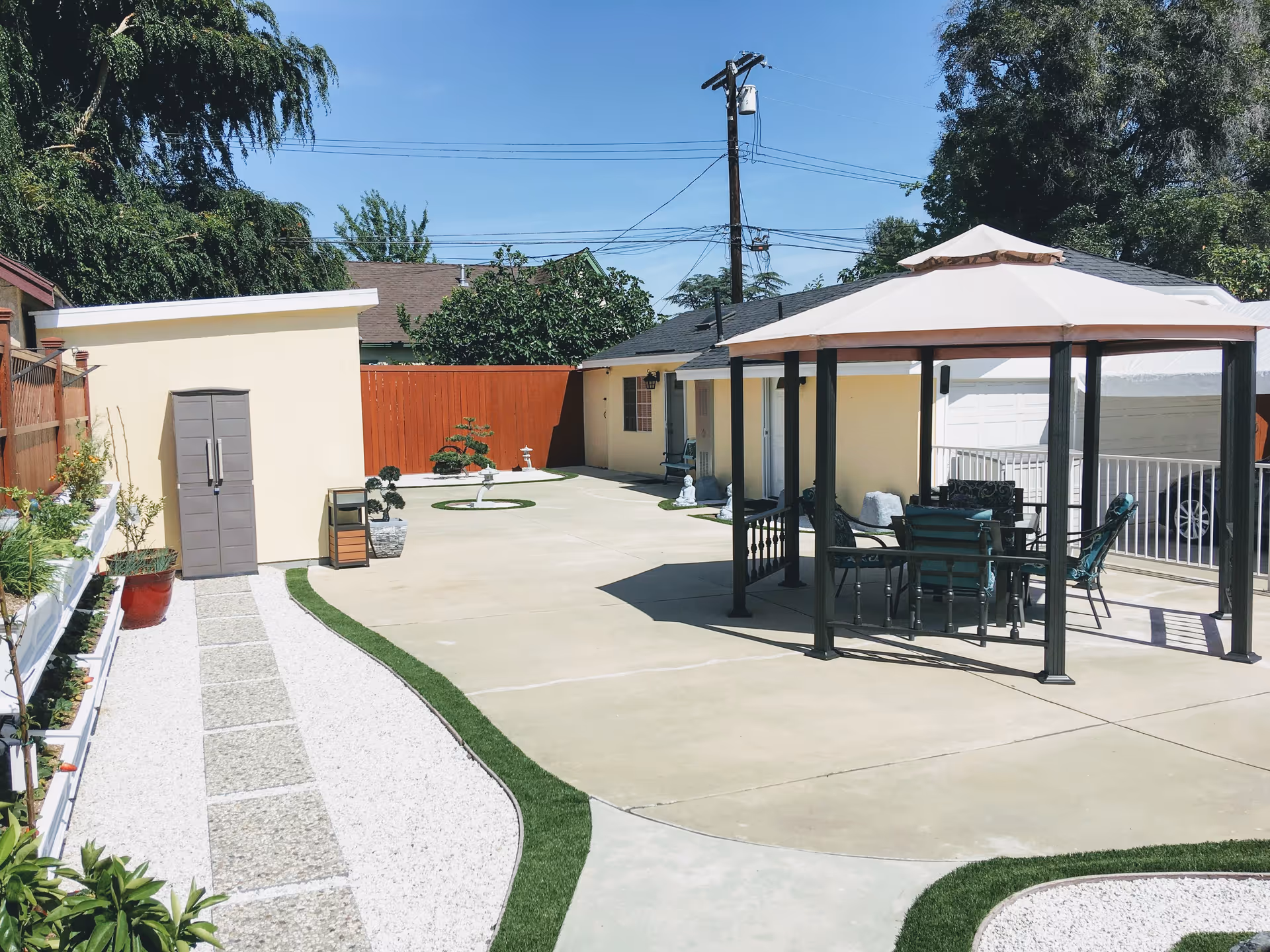 Outdoor patio area with a gazebo featuring a table and chairs, surrounded by a concrete surface and landscaped with small plants and decorative gravel. There is a small shed and a wooden fence in the background under a clear blue sky.