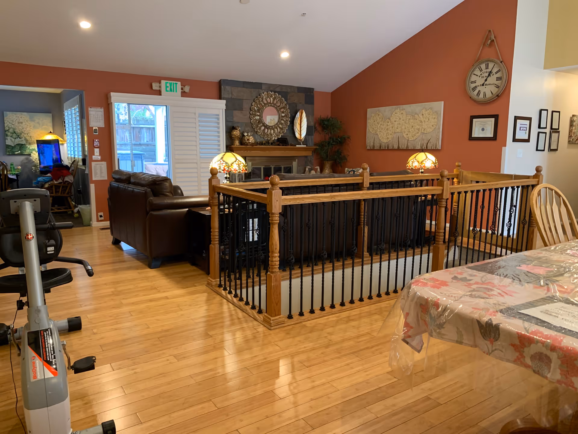 Interior view of a senior living facility showing a cozy living room area with brown leather sofas, a fireplace with decorative items above it, and a wooden railing surrounding a stairwell. The walls are painted a warm terracotta color and decorated with a large clock and framed artwork. To the right, there is a dining table covered with a floral tablecloth and a plastic cover. On the left, there is an exercise bike and a glimpse into another room with a blue wall and a lamp.