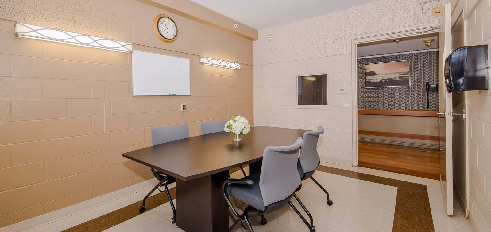 A small conference or meeting room with a rectangular dark wood table surrounded by four gray office chairs on wheels. The walls are beige with two modern wall lights, a clock, and a whiteboard mounted on one wall. A vase with white flowers is placed in the center of the table. An open door reveals a hallway with wooden flooring and a framed picture on the wall.
