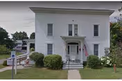 Two-story white building with a small porch, shrubs, and an American flag