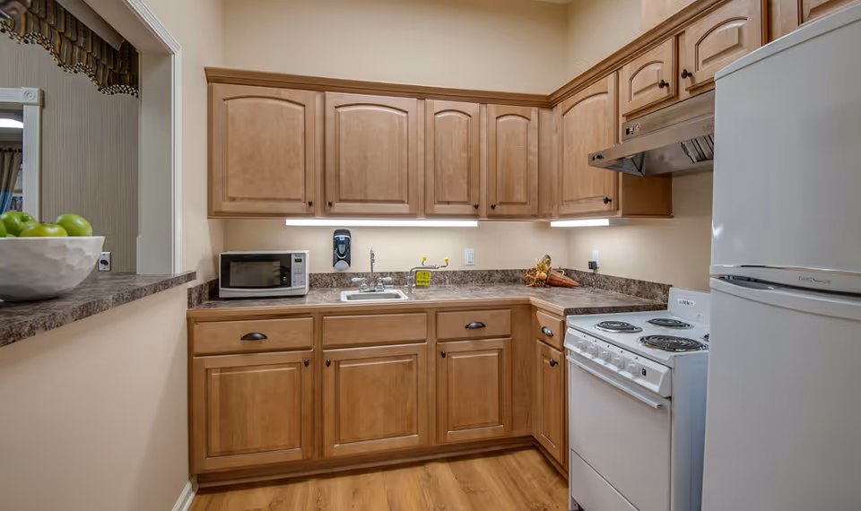 A compact kitchen with wooden cabinets, a white electric stove with four burners, a white refrigerator, a microwave on the countertop, a small sink, and under-cabinet lighting. There is a bowl of green apples on a raised counter to the left.