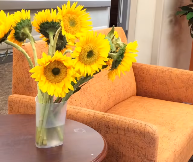 A clear vase with a bouquet of bright yellow sunflowers placed on a round wooden table in front of an orange upholstered armchair in a cozy indoor setting.
