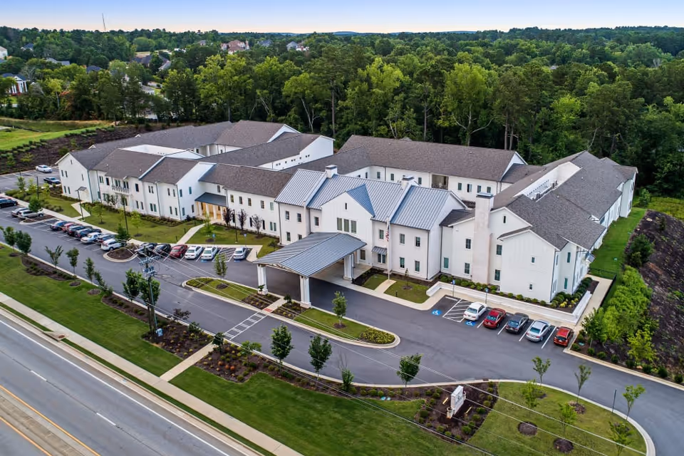 Aerial view of The Claiborne at Westlake, a large two-story senior living facility surrounded by greenery and trees. The building has a light-colored exterior with a covered entrance and multiple parking spaces in front. The facility is bordered by a road and landscaped areas with small trees and shrubs.