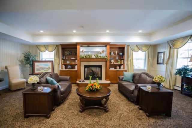 A cozy living room with two brown leather sofas facing each other, each with a side table holding a vase of flowers. A round wooden coffee table with a floral centerpiece is in the middle. Behind the coffee table is a fireplace with a wooden mantel and built-in shelves on either side, decorated with books and ornaments. The room has large windows with yellow curtains and patterned carpet flooring.