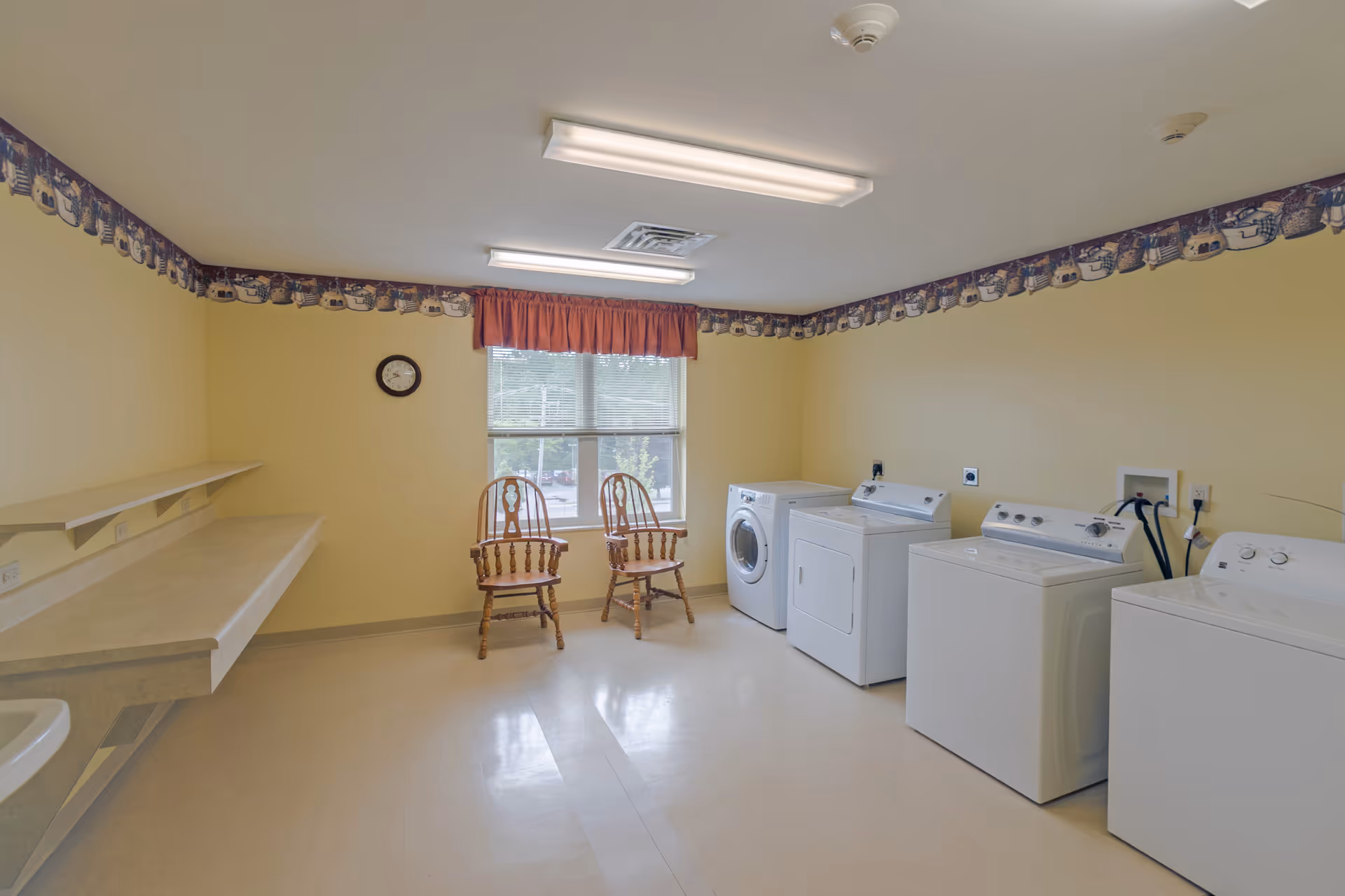A laundry room with four white washing machines and dryers lined up against a yellow wall. Two wooden chairs are placed near a window with blinds and a red valance. The room has beige flooring, a clock on the wall, fluorescent ceiling lights, and a decorative border near the ceiling featuring a pattern of teapots and cups.