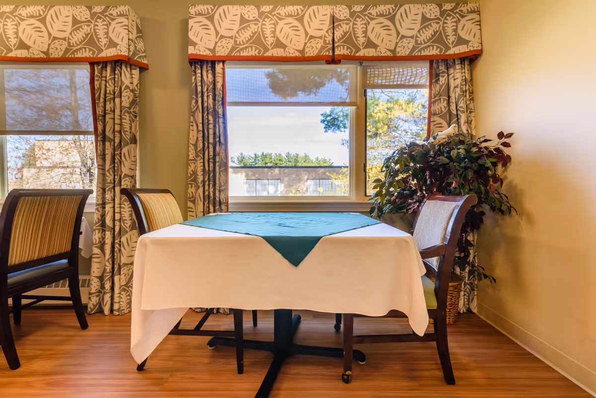 A dining area with a table covered by a white tablecloth and a green table runner, surrounded by wooden chairs with striped cushions. The room has large windows with patterned curtains and valances, allowing natural light to brighten the space. A potted plant is placed in the corner near the window.
